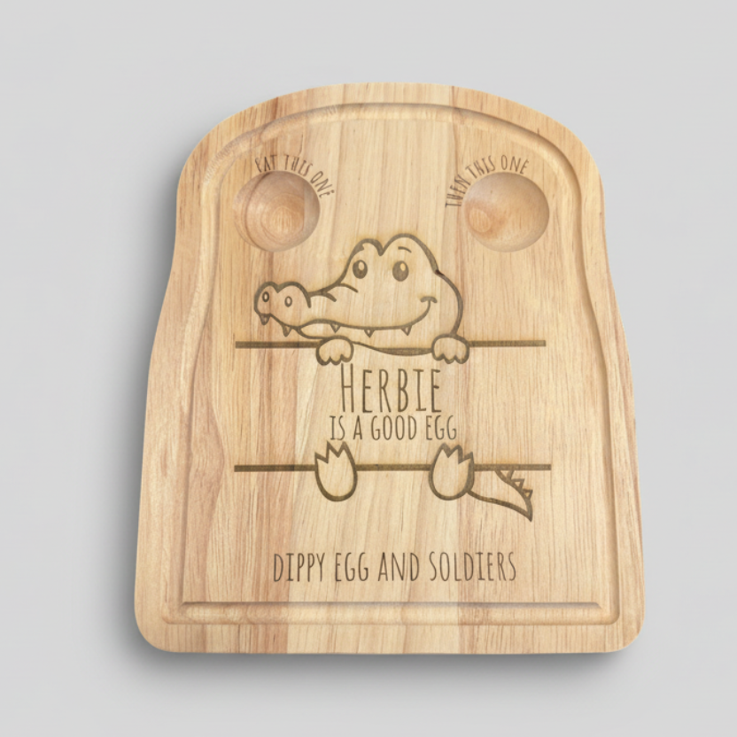Wooden cutting board with engraved text and a cartoon crocodile design, surrounded by eggs on a neutral background.