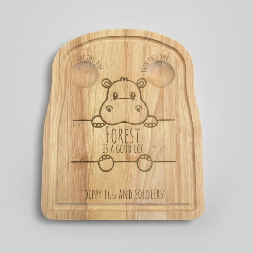 Wooden cutting board with a hippo design and text on a table with eggs and bread.
