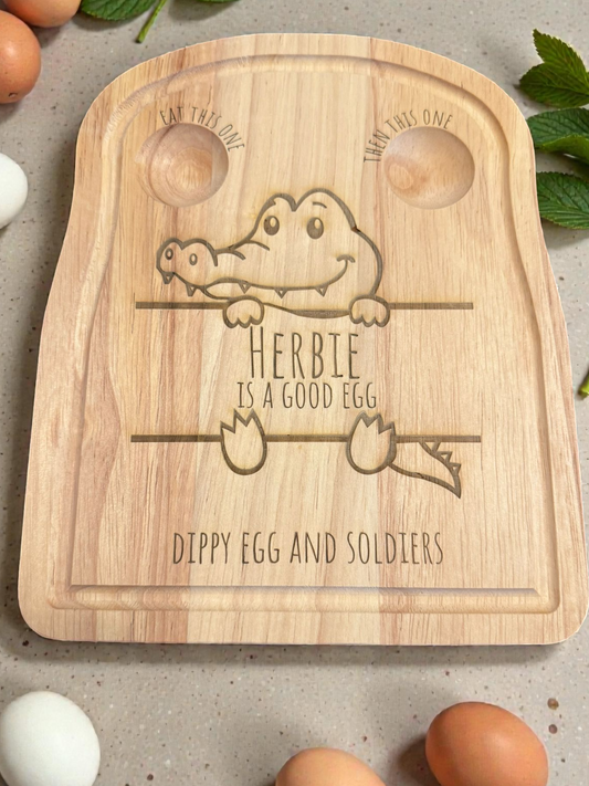 Wooden cutting board with engraved text and a cartoon crocodile design, surrounded by eggs on a neutral background.