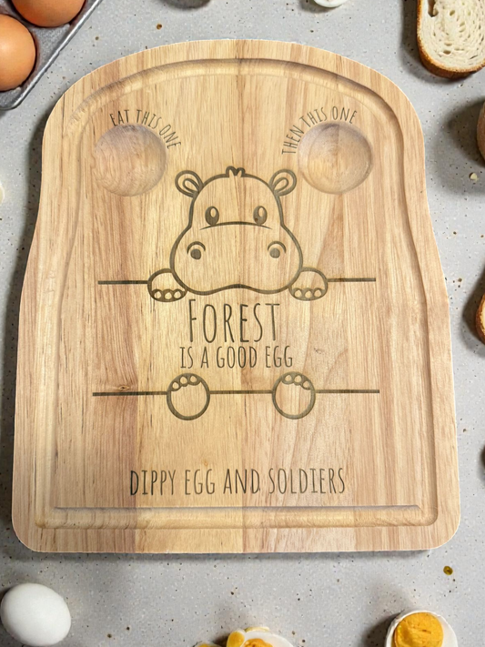 Wooden cutting board with a hippo design and text on a table with eggs and bread.